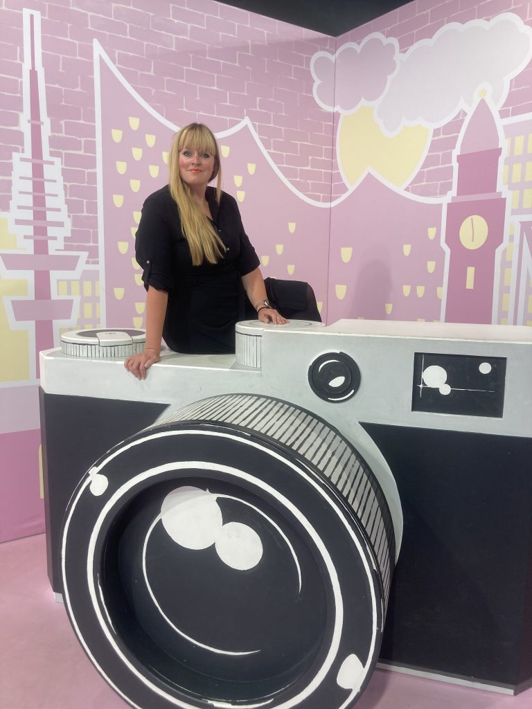 Daniela Friedrich sietzt vor einem rosa Hintergrund auf einer übergroßen Kamera